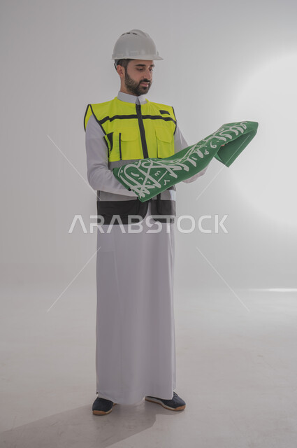 المشاركة بالاحتفال بالمناسبات والأعياد الوطنية، إحياء ذكرى اليوم الوطني السعودي، بورتريه لمهندس عربي خليجي سعودي يرتدي الثوب التقليدي والسترة وقبعة الحماية ويحمل راية المملكة العربية السعودية بين يديه بإيماءات الفخر والاعتزاز، الجسم بالطول الكامل، خلفية رمادية