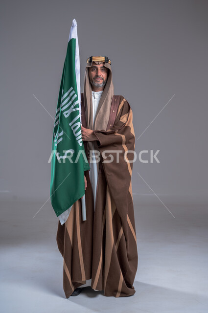 مفهوم الاصالة والعراقة عند العرب، بورتريه لرجل عربي خليجي سعودي يرتدي العقال الذهبي والبشت يحمل بيده علم التأسيس، يوم بدينا 22 فبراير, إحياء تراث المملكة, الاحتفالات بالمناسبات الوطنية, ذكرى تأسيس الدولة الأولى 1727م، الوقوف باستقامة وشموخ، خلفية رمادية