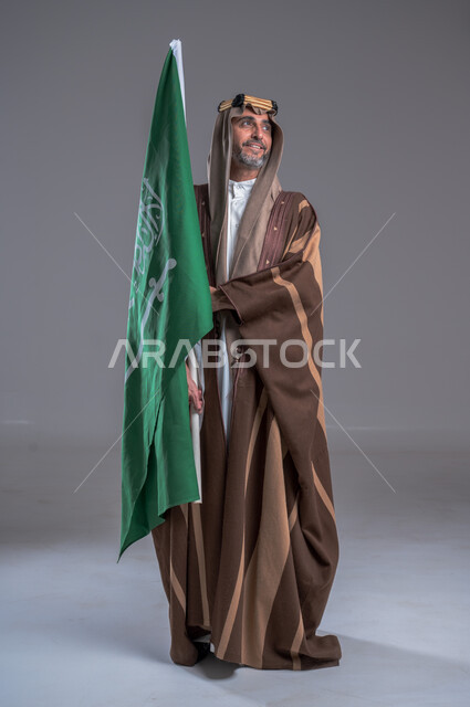 مفهوم الاصالة والعراقة عند العرب، بورتريه لرجل عربي خليجي سعودي يرتدي العقال الذهبي والبشت يحمل بيده علم التأسيس، يوم بدينا 22 فبراير, إحياء تراث المملكة, الاحتفالات بالمناسبات الوطنية, ذكرى تأسيس الدولة الأولى 1727م، الوقوف باستقامة وشموخ، خلفية رمادية