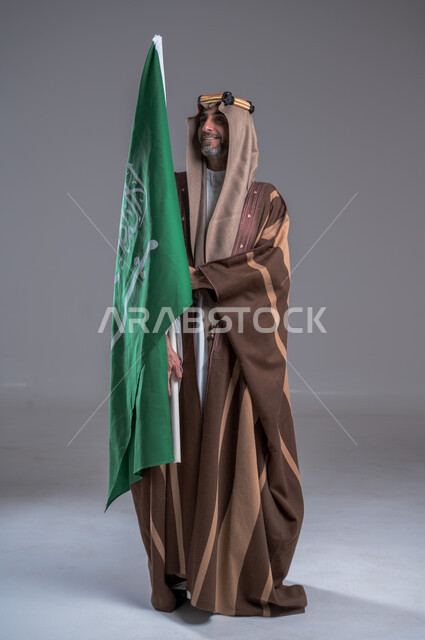 الوقوف باستقامة وشموخ، مفهوم الاصالة والعراقة عند العرب، بورتريه لرجل عربي خليجي سعودي يرتدي العقال الذهبي والبشت يحمل بيده علم التأسيس، يوم بدينا 22 فبراير, إحياء تراث المملكة, الاحتفالات بالمناسبات الوطنية, ذكرى تأسيس الدولة الأولى 1727م، خلفية رمادية
