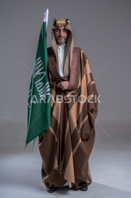 الوقوف باستقامة وشموخ، مفهوم الاصالة والعراقة عند العرب، بورتريه لرجل عربي خليجي سعودي يرتدي العقال الذهبي والبشت يحمل بيده علم التأسيس، يوم بدينا 22 فبراير, إحياء تراث المملكة, الاحتفالات بالمناسبات الوطنية, ذكرى تأسيس الدولة الأولى 1727م، خلفية رمادية