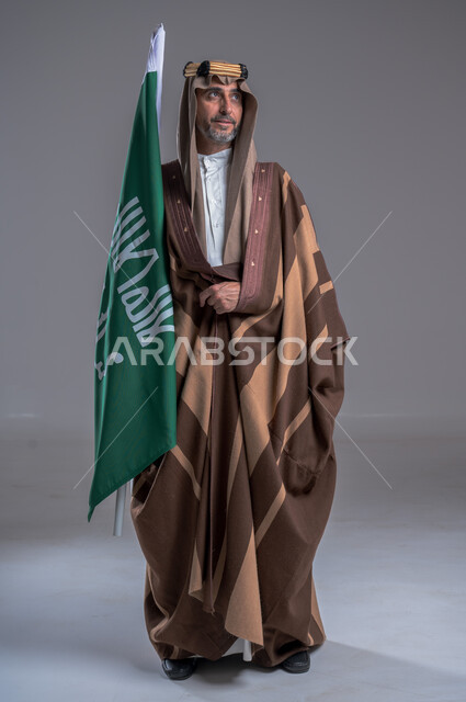 ذكرى تأسيس الدولة السعودية الأولى 22 فبراير، الفخر والاعتزاز بإنجازات الوطن، الاحتفال بالمناسبات الوطنية, شعار الهوية الوطنية وراية الإسلام والتوحيد، يوم بدينا 1727 م، بورتريه لرجل عربي خليجي سعودي يرتدي العقال الذهبي والبشت يحمل بيده علم التأسيس, خلفية رمادية