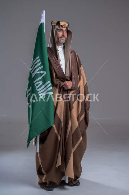 The anniversary of the founding of the first Saudi state, February 22, pride and honor in the achievements of the nation, celebrating national occasions, the slogan of national identity and the banner of Islam and monotheism, the day we started 1727 AD, a portrait of a Saudi Gulf Arab man wearing a golden agal and a bisht holding the founding flag in his hand, gray background