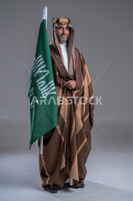 شعار الهوية الوطنية وراية الإسلام والتوحيد، يوم بدينا 1727 م، بورتريه لرجل عربي خليجي سعودي يرتدي العقال الذهبي والبشت يحمل بيده علم التأسيس, ذكرى تأسيس الدولة السعودية الأولى 22 فبراير، الفخر والاعتزاز بإنجازات الوطن، الاحتفال بالمناسبات الوطنية, خلفية رمادية