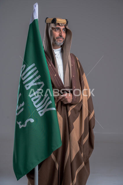 شعار الهوية الوطنية وراية الإسلام والتوحيد، يوم بدينا 1727 م، بورتريه لرجل عربي خليجي سعودي يرتدي العقال الذهبي والبشت يحمل بيده علم التأسيس, ذكرى تأسيس الدولة السعودية الأولى 22 فبراير، الفخر والاعتزاز بإنجازات الوطن، الاحتفال بالمناسبات الوطنية, خلفية رمادية
