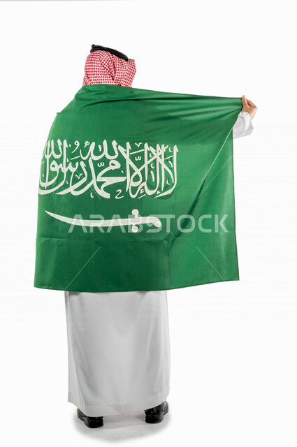 تصوير من الخلف لرجل عربي خليجي سعودي يضع على ظهره علم المملكة العربية السعودية ، ذكرى اليوم الوطني السعودي ،الاحتفال بالعيد الوطني السعودي