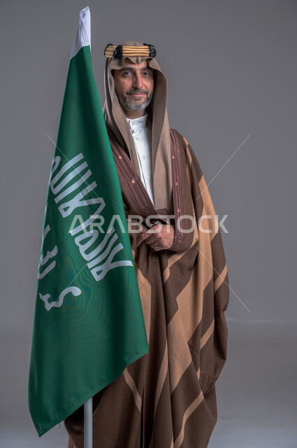 الفخر والاعتزاز بإنجازات الوطن، شعار الهوية الوطنية وراية الإسلام والتوحيد، يوم بدينا 1727 م، بورتريه لرجل عربي خليجي سعودي مبتسم يرتدي العقال الذهبي والبشت يحمل بيده علم التأسيس, ذكرى تأسيس الدولة السعودية الأولى 22 فبراير، خلفية رمادية