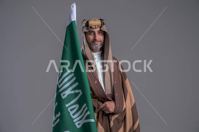 الفخر والاعتزاز بإنجازات الوطن، شعار الهوية الوطنية وراية الإسلام والتوحيد، يوم بدينا 1727 م، بورتريه لرجل عربي خليجي سعودي مبتسم يرتدي العقال الذهبي والبشت يحمل بيده علم التأسيس, ذكرى تأسيس الدولة السعودية الأولى 22 فبراير، خلفية رمادية