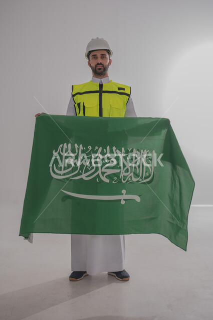 Raising the flag of the Kingdom of Saudi Arabia with gestures of pride and honor, participating in the celebration of national occasions and holidays, commemorating the Saudi National Day, portrait of a Saudi Arabian Gulf engineer wearing a jacket and a protective hat standing and looking at the camera with pleasure, full body length, gray background