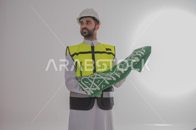 رفع راية المملكة العربية السعودية بإيماءات الفخر والاعتزاز، المشاركة بالاحتفال بالمناسبات والأعياد الوطنية، إحياء ذكرى اليوم الوطني السعودي ٢٣ سبتمبر، بورتريه لمهندس عربي خليجي سعودي يرتدي السترة وقبعة الحماية يقف وينظر لمكان ما بعيد، خلفية رمادية