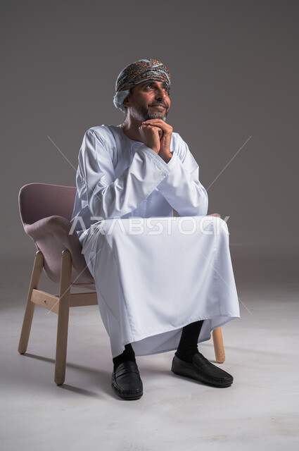 Looking at something with expressions of thinking and absent-mindedness, portrait of an Arab Gulf Omani man wearing a dishdasha and turban sitting on a comfortable chair and putting his hands under his chin, contemplation and planning for the future, finding a solution to a problem and making decisions, gray background