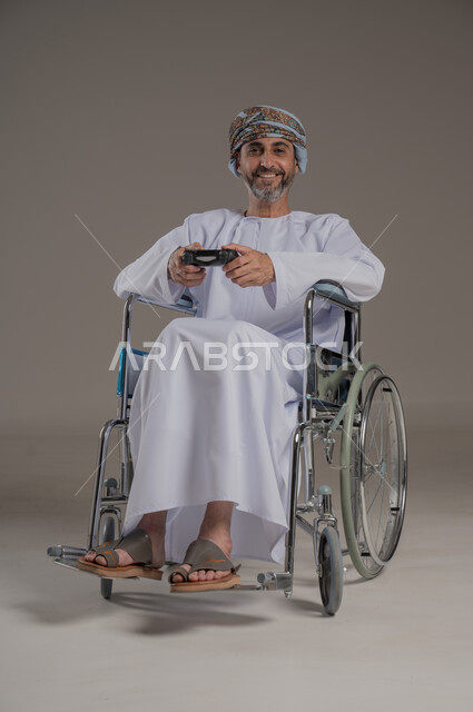 تجربة مثيرة في الأجهزة الحديثة الالكترونية، نشاطات ترفيهية لذوي الهمم، التفاعل والاندماج مع ألعاب البلايستيشن, بورتريه لرجل عربي خليجي عماني مبتسم مقعد يرتدي العمامة والدشداشة يجلس على كرسي متحرك يحمل بيده عصا التحكم، النظر الى الكاميرا بتعابير الفرح، خلفية رمادية