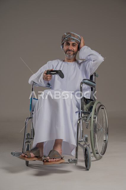 تجربة مثيرة في الأجهزة الحديثة الالكترونية، نشاطات ترفيهية لذوي الهمم، التفاعل والاندماج مع ألعاب البلايستيشن, بورتريه لرجل عربي خليجي عماني مبتسم مقعد يرتدي العمامة والدشداشة يجلس على كرسي متحرك يحمل بيده عصا التحكم، النظر الى الكاميرا بتعابير الفرح، خلفية رمادية