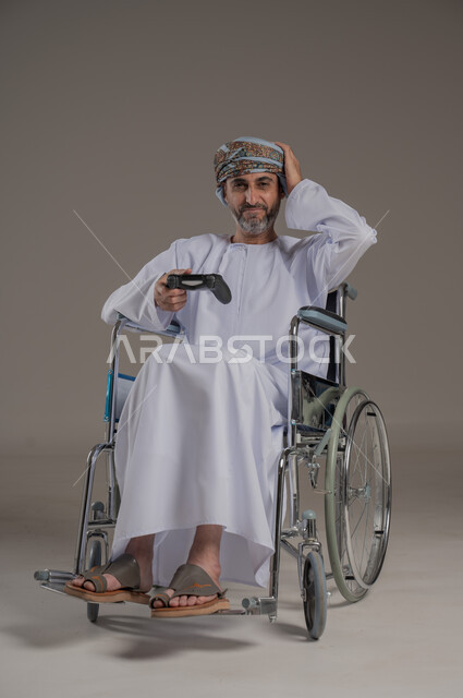 تجربة مثيرة في الأجهزة الحديثة الالكترونية، نشاطات ترفيهية لذوي الهمم، التفاعل والاندماج مع ألعاب البلايستيشن, بورتريه لرجل عربي خليجي عماني مبتسم مقعد يرتدي العمامة والدشداشة يجلس على كرسي متحرك يحمل بيده عصا التحكم، النظر الى الكاميرا بتعابير الفرح، خلفية رمادية
