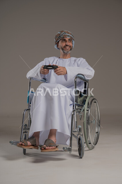 نشاطات ترفيهية لذوي الهمم، التفاعل والاندماج مع ألعاب البلايستيشن, تجربة مثيرة في الأجهزة الحديثة الالكترونية، بورتريه لرجل عربي خليجي عماني مبتسم مقعد يرتدي العمامة والدشداشة يجلس على كرسي متحرك يحمل بيده قبضة التحكم، النظر الى مكان ما بإيماءات الفوز والانتصار، خلفية رمادية