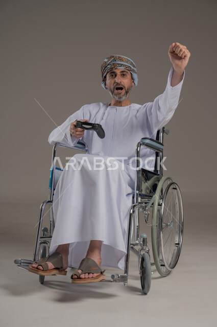  نشاطات ترفيهية لذوي الهمم، التفاعل والاندماج مع ألعاب البلايستيشن, تجربة مثيرة في الأجهزة الحديثة الالكترونية، بورتريه لرجل عربي خليجي عماني مبتسم مقعد يرتدي العمامة والدشداشة يجلس على كرسي متحرك يحمل بيده قبضة التحكم، النظر الى مكان ما بإيماءات الفوز والانتصار، خلفية رمادية
