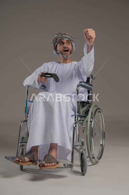  نشاطات ترفيهية لذوي الهمم، التفاعل والاندماج مع ألعاب البلايستيشن, تجربة مثيرة في الأجهزة الحديثة الالكترونية، بورتريه لرجل عربي خليجي عماني مبتسم مقعد يرتدي العمامة والدشداشة يجلس على كرسي متحرك يحمل بيده قبضة التحكم، النظر الى مكان ما بإيماءات الفوز والانتصار، خلفية رمادية