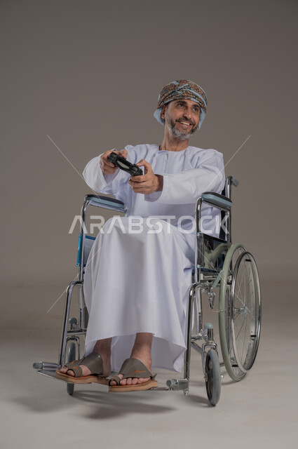  نشاطات ترفيهية لذوي الهمم، التفاعل والاندماج مع ألعاب البلايستيشن, تجربة مثيرة في الأجهزة الحديثة الالكترونية، بورتريه لرجل عربي خليجي عماني مبتسم مقعد يرتدي العمامة والدشداشة يجلس على كرسي متحرك يحمل بيده قبضة التحكم، النظر الى مكان ما بإيماءات الفوز والانتصار، خلفية رمادية