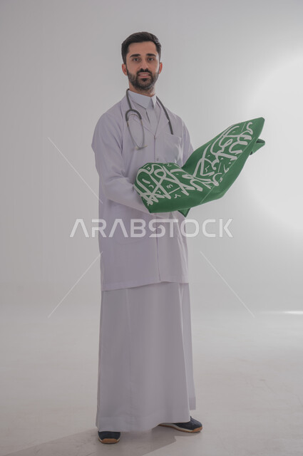 إيماءات الفخر والاعتزاز برفع راية المملكة العربية السعودية، المشاركة بالاحتفال بالمناسبات والأعياد الوطنية، إحياء ذكرى اليوم الوطني السعودي٢٣ سبتمبر، بورتريه لطبيب عربي خليجي سعودي يرتدي المعطف الطبي ويضع سماعة الفحص حول عنقه ينظر للكاميرا بسرور، خلفية رمادية