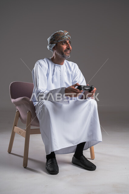 Victory and winning gestures, interaction and integration with PlayStation games, entertainment electronic technologies, portrait of an Arab Gulf Omani man wearing a turban and dishdasha sitting on a comfortable chair holding a joystick in his hand, a fun and exciting entertainment experience on modern devices, gray background