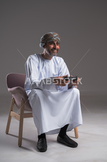 ايماءات الانتصار والفوز، التفاعل والاندماج مع ألعاب البلايستيشن، تقنيات الكترونية ترفيهيه, بورتريه لرجل عربي خليجي عماني يرتدي العمامة والدشداشة يجلس على كرسي مريح يحمل بيده عصا التحكم، تجربة ترفيهية ممتعة ومثيرة في الأجهزة الحديثة، خلفية رمادية