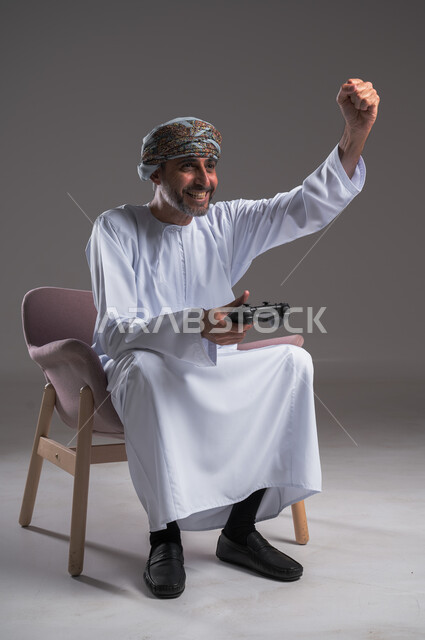 ايماءات الانتصار والفوز، التفاعل والاندماج مع ألعاب البلايستيشن، تقنيات الكترونية ترفيهيه, بورتريه لرجل عربي خليجي عماني يرتدي العمامة والدشداشة يجلس على كرسي مريح يحمل بيده عصا التحكم، تجربة ترفيهية ممتعة ومثيرة في الأجهزة الحديثة، خلفية رمادية