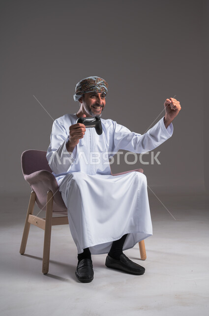 ايماءات الانتصار والفوز، التفاعل والاندماج مع ألعاب البلايستيشن، تقنيات الكترونية ترفيهيه, بورتريه لرجل عربي خليجي عماني يرتدي العمامة والدشداشة يجلس على كرسي مريح يحمل بيده عصا التحكم، تجربة ترفيهية ممتعة ومثيرة في الأجهزة الحديثة، خلفية رمادية