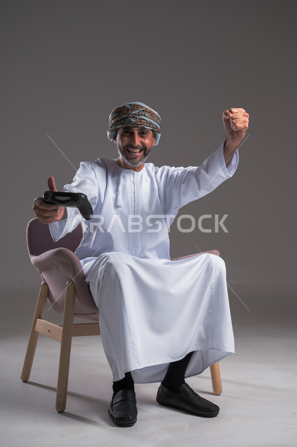 ايماءات الانتصار والفوز، التفاعل والاندماج مع ألعاب البلايستيشن، تقنيات الكترونية ترفيهيه, بورتريه لرجل عربي خليجي عماني يرتدي العمامة والدشداشة يجلس على كرسي مريح يحمل بيده عصا التحكم، تجربة ترفيهية ممتعة ومثيرة في الأجهزة الحديثة، خلفية رمادية