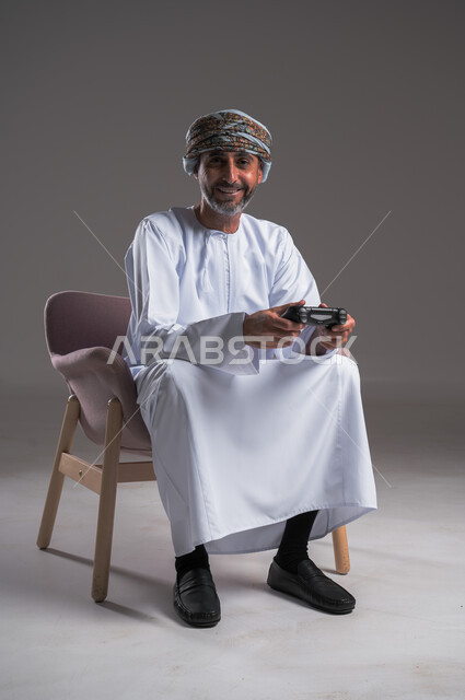 التفاعل والاندماج مع ألعاب البلايستيشن، تقنيات الكترونية ترفيهيه, بورتريه لرجل عربي خليجي عماني يرتدي العمامة والدشداشة يجلس على كرسي مريح يحمل بيده قبضة التحكم، تجربة ترفيهية ممتعة ومثيرة في الأجهزة الحديثة، النظر الى الكاميرا بإيماءات الفرح، صورة لكامل الجسم، خلفية رمادية