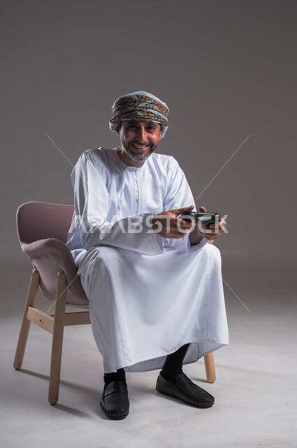 Interaction and integration with PlayStation games, entertainment electronic technologies, portrait of an Arab Gulf Omani man wearing a turban and a dishdasha sitting on a comfortable chair holding a control handle in his hand, a fun and exciting entertainment experience in modern devices, looking at the camera with gestures of joy, a full body image, a gray background