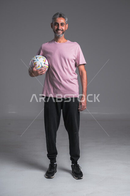 Professional team sports, practicing the hobby of playing football, close-up portrait of a Saudi Arabian Gulf man wearing goalkeeper gloves holding the ball in his hand and looking at the camera with a smile, full body photo, gray background