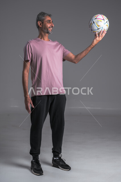 Professional team sports, practicing the hobby of playing football, a close-up portrait of a Saudi Arabian Gulf man wearing sportswear and gloves holding a ball in his hand and looking at it, full body photo, gray background