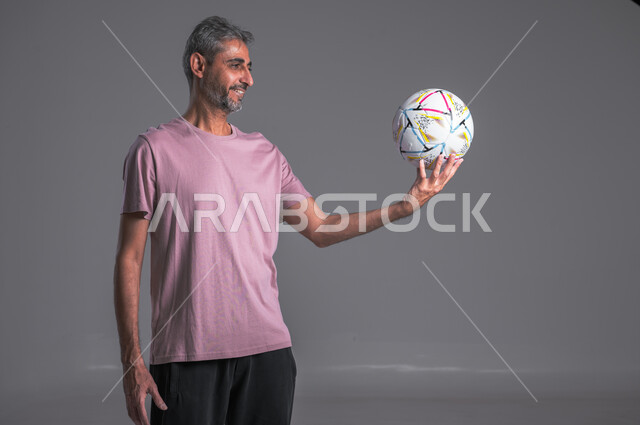 احتراف الرياضات الجماعية، ممارسة هواية اللعب بكرة القدم،  بورتريه مقرب لرجل عربي سعودي خليجي ويرتدي قفازات حراسة المرمى  يحمل الكره بيده وينظر اليها  ، صوره لكامل الجسم ، خلفية رمادية