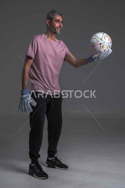 هواية اللعب والتسليه بكره القدم ،ممارسة تمرينات رياضة ، بورتريه لرجل عربي خليجي سعودي ويرتدي قفازات حراسة المرمى يحمل بيده الكرة وينظر اليها ،  صوره للجسم بالطول الكامل ، خلفيه رماديه