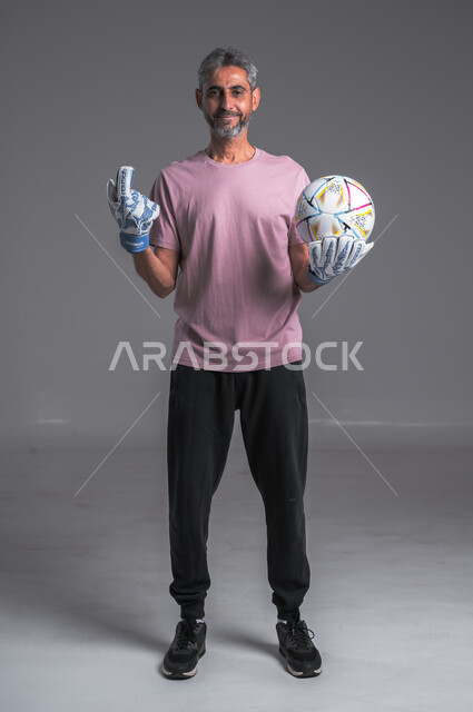 الحصول على المركز الاول ، هواية اللعب والتسليه بكره القدم ، ممارسة تمرينات رياضة ، بورتريه لرجل عربي خليجي سعودي ويرتدي قفازات حراسة المرمى ينظر الى الكاميرا يرفع اصبع السبابة للأعلى، صوره للجسم بالطول الكامل ، خلفيه رماديه