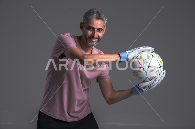 هواية اللعب والتسليه بكره القدم ، ممارسة تمرينات رياضة ، بورتريه لرجل عربي خليجي سعودي يرتدي قفازات حراسة المرمى وينظر الى الكاميرا بثقة يحمل بيديه الكرة ، خلفيه رماديه