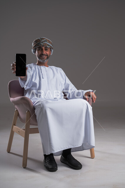 Using modern technology, displaying a blank black screen on a mobile phone, looking at the camera with gestures of joy and happiness, portrait of a smiling Omani Gulf Arab man wearing a turban and a dishdasha sitting on a comfortable chair, holding a mobile phone and pointing to it, full body image, gray background