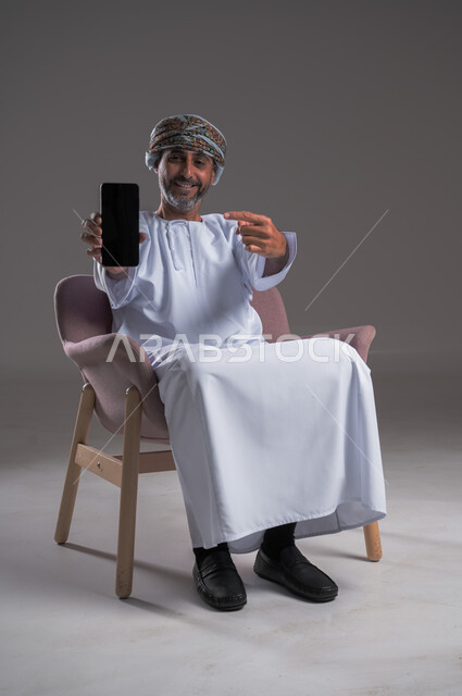 Using modern technology, displaying a blank black screen on a mobile phone, looking at the camera with gestures of joy and happiness, portrait of a smiling Omani Gulf Arab man wearing a turban and a dishdasha sitting on a comfortable chair, holding a mobile phone and pointing to it, full body image, gray background