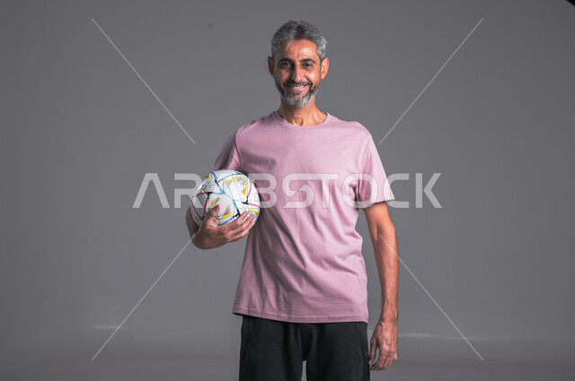 Professional team sports, practicing the hobby of playing football, close-up portrait of a Saudi Arabian Gulf man wearing goalkeeper gloves, holding the ball in his hand and looking at the camera with a smile, gray background