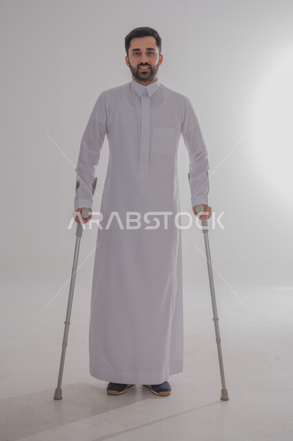 أسلوب حياة ذوي الهمم، استخدام العكازات الطبية للمساعدة على الحركة والتنقل، تحسين جودة حياة ذوي الاحتياجات الخاصة ودمجهم في المجتمع، بورتريه لشاب عربي خليجي سعودي يرتدي الثوب التقليدي يقف ويستند على عصا حديدية، الجسم بالطول الكامل، خلفية رمادية