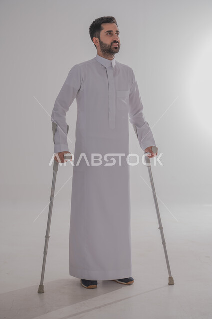 أسلوب حياة ذوي الهمم، استخدام العكازات الطبية للمساعدة على الحركة والتنقل، تحسين جودة حياة ذوي الاحتياجات الخاصة ودمجهم في المجتمع، بورتريه لشاب عربي خليجي سعودي يرتدي الثوب التقليدي يقف ويستند على عصا حديدية، الجسم بالطول الكامل، خلفية رمادية
