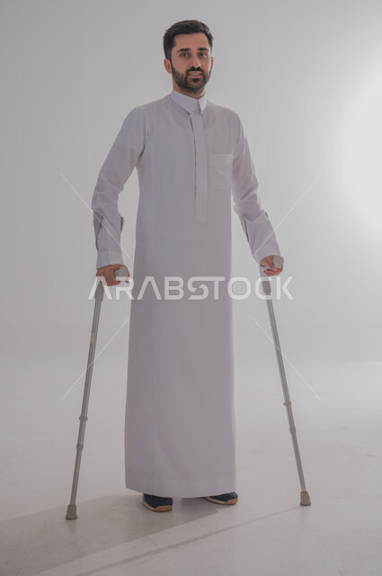 أسلوب حياة ذوي الهمم، استخدام العكازات الطبية للمساعدة على الحركة والتنقل، تحسين جودة حياة ذوي الاحتياجات الخاصة ودمجهم في المجتمع، بورتريه لشاب عربي خليجي سعودي يرتدي الثوب التقليدي يقف ويستند على عصا حديدية، الجسم بالطول الكامل، خلفية رمادية