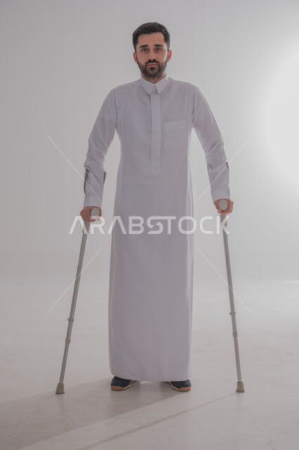 أسلوب حياة ذوي الهمم، استخدام العكازات الطبية للمساعدة على الحركة والتنقل، تحسين جودة حياة ذوي الاحتياجات الخاصة ودمجهم في المجتمع، بورتريه لشاب عربي خليجي سعودي يرتدي الثوب التقليدي يقف ويستند على عصا حديدية، الجسم بالطول الكامل، خلفية رمادية