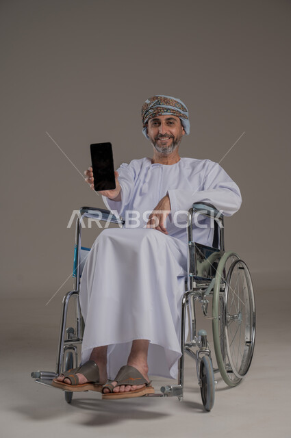 Using modern technology, displaying a blank black screen on a mobile phone, looking at the camera with gestures of joy and happiness, portrait of a smiling Omani Gulf Arab man with special needs wearing a turban and dishdasha sitting in a wheelchair holding a mobile phone, full body image, gray background