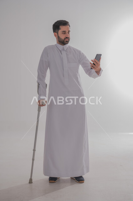 تحسين جودة حياة ذوي الاحتياجات الخاصة باستخدام التقنيات والتكنولوجيا، الاعتماد على العكازات الطبية للمساعدة على الحركة، بورتريه لشاب عربي خليجي سعودي يرتدي الثوب التقليدي يقف ويستند على عصا حديدية يستخدم جهاز الهاتف المحمول، الجسم بالطول الكامل، خلفية رمادية