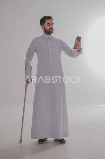 تحسين جودة حياة ذوي الاحتياجات الخاصة باستخدام التقنيات والتكنولوجيا، الاعتماد على العكازات الطبية للمساعدة على الحركة، بورتريه لشاب عربي خليجي سعودي يرتدي الثوب التقليدي يقف ويستند على عصا حديدية يستخدم جهاز الهاتف المحمول، الجسم بالطول الكامل، خلفية رمادية