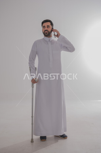 الاعتماد على العكازات الطبية للمساعدة على الحركة، تحسين جودة حياة ذوي الاحتياجات الخاصة باستخدام التقنيات والتكنولوجيا، بورتريه لشاب عربي خليجي سعودي يرتدي الثوب التقليدي يقف ويستند على عصا حديدية يجري مكالمة هاتفية عبر الجوال، الجسم بالطول الكامل، خلفية رمادية