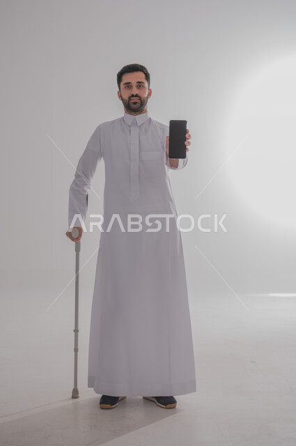 الاعتماد على العكازات الطبية للمساعدة على الحركة، تحسين جودة حياة ذوي الاحتياجات الخاصة باستخدام التقنيات والتكنولوجيا، بورتريه لشاب عربي خليجي سعودي يرتدي الثوب التقليدي يقف ويستند على عصا حديدية يعرض شاشة جوال سوداء فارغة، الجسم بالطول الكامل، خلفية رمادية