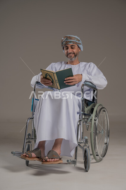 مفهوم قراءة القران وحفظه وتجويده، ذوي الإحتياجات الخاصة والهمم, بورتريه لشاب عربي خليجي عماني مبتسم يرتدي العمامة والدشداشة يجلس على كرسي متحرك ويحمل بيديه المصحف الشريف، خلفية رمادي