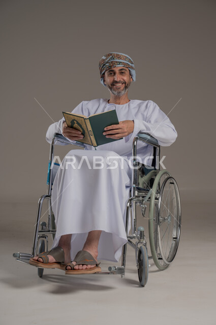 مفهوم قراءة القران وحفظه وتجويده، ذوي الإحتياجات الخاصة والهمم, بورتريه لشاب عربي خليجي عماني مبتسم يرتدي العمامة والدشداشة يجلس على كرسي متحرك ويحمل بيديه المصحف الشريف، خلفية رمادي